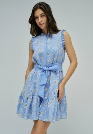 Mujer vistiendo un vestido sin mangas azul claro con estampado floral, volantes, botones en la parte delantera y cinturón atado en la cintura, posando contra un fondo liso.