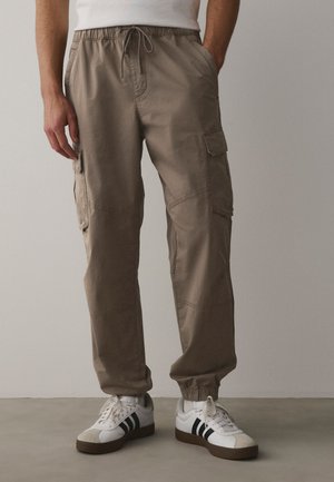 Uomo che indossa pantaloni cargo beige con polsini elastici e sneakers bianche con strisce nere, in piedi su uno sfondo semplice.