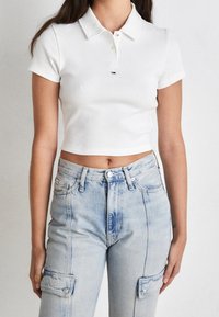 Kvinna som bär en vit kortärmad polotröja och högmidjade ljusblå denim cargobyxor, stående mot en neutral bakgrund.