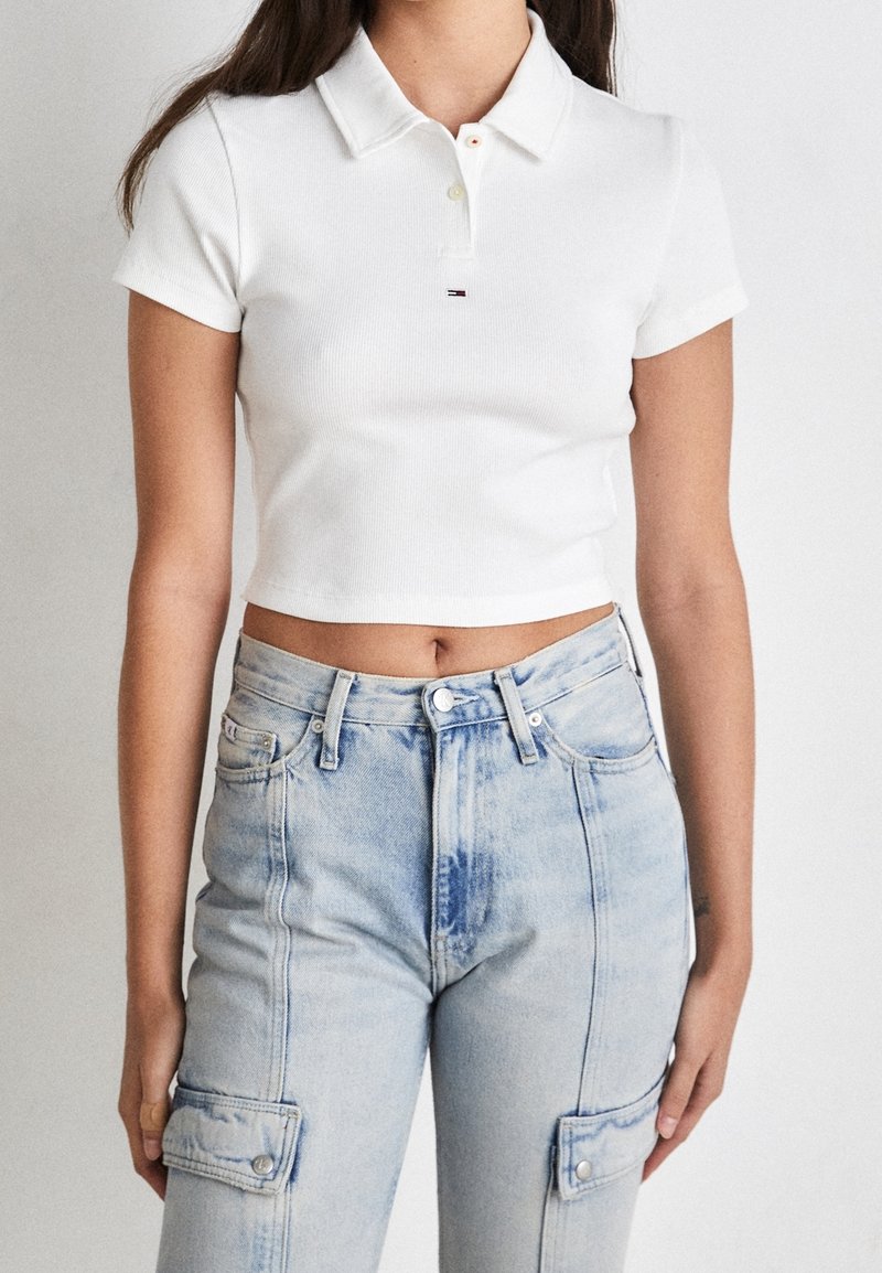 Kvinna som bär en vit kortärmad polotröja och högmidjade ljusblå denim cargobyxor, stående mot en neutral bakgrund.