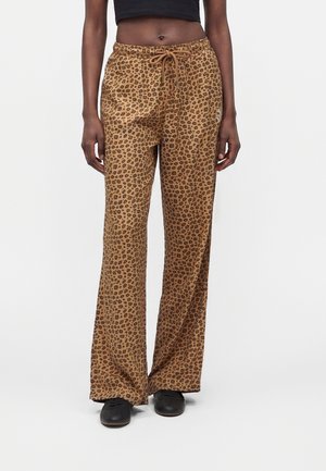 Personne portant un pantalon large imprimé léopard marron avec une taille à cordon et des chaussures noires sur un fond uni.