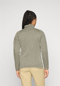 Woman with braided hair wears a gray fleece jacket and beige pants, standing against a plain light background, facing away from the camera.