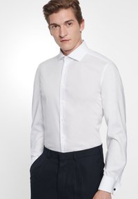 Vit skjorta med långa ärmar, knapp-down krage och textilstruktur; matchad med mörka skräddarsydda byxor och manschetter.