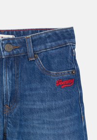 Pantalones vaqueros de mezclilla azul con costuras en contraste, diseño de cinco bolsillos, cierre de botón metálico y logotipo de la marca bordado en rojo en el bolsillo frontal.