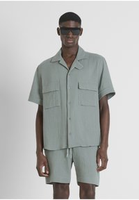 Lichtgroene, gestructureerde kortekraagshirt met twee voorzakken, gecombineerd met bijpassende shorts. Model draagt een zwarte zonnebril.