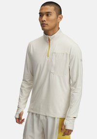 Camicia a maniche lunghe di un leggero colore crema, con una zip frontale a metà. Presenta una tasca sul petto e aperture per il pollice nelle maniche per una maggiore copertura.