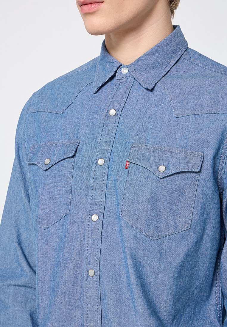 Camisa de mezclilla azul claro con cuello abotonado, botones a presión plateados, dos bolsillos en el pecho y costuras tonales. Textura suave y corte clásico.