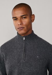 Pull zipé gris charbon avec col et poignets côtelés, fabriqué en tissu tricoté texturé. Présente un petit logo sur la poitrine.