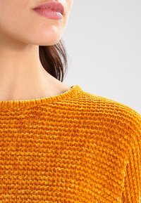 Leuchtend orangefarbener, strukturierter Pullover mit rundem Ausschnitt. Der Stoff wirkt weich und plüschig und zeigt ein durchgängiges Webmuster.