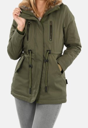 Persoon draagt een olijfgroene parka met een capuchon met bontvoering, borstzakken met rits, een taillekoord en voorzakken met klep, gecombineerd met grijze broek.