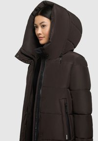Doudoune marron foncé avec une grande capuche, fermeture éclair frontale et poches latérales. Le matériau semble lisse avec un design matelassé et peu d'accents.