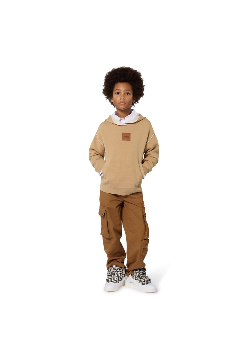 Felpa cappuccio beige con un'etichetta in pelle, indossata sopra una camicia bianca; abbinata a pantaloni cargo marrone e sneakers bianche con un design intrecciato nero.