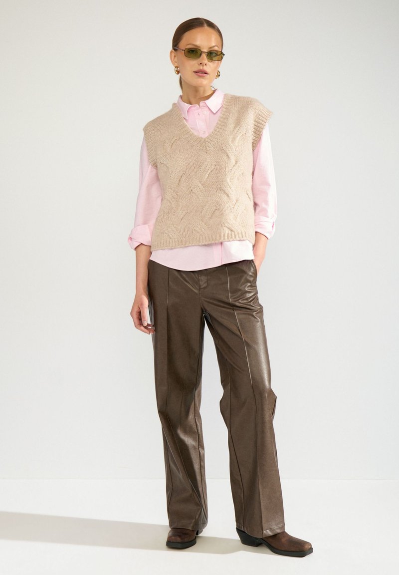 Femme portant un gilet en maille beige sur une chemise rose, un pantalon en cuir marron, des chaussures marron, des lunettes de soleil teintées de vert et des boucles d'oreilles créoles dorées, debout sur fond blanc.