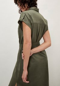 Olivenfarbenes Hemdkleid mit kurz gerollten Ärmeln, tailliertem Bund und seitlichen Schlitzen. Weicher Stoff mit subtiler Textur und zwei Brusttaschen.