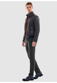 Zwarte gewatteerde leren jas, oranje trui, grijze geblokte broek en navy schoenen met een band. Model staat zijwaarts tegen een neutrale achtergrond.