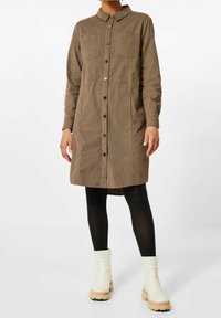 Vestito camicia marrone con bottoni, due tasche sul petto, maniche lunghe e tessuto testurizzato, abbinato a leggings neri e stivaletti bianchi alla caviglia.