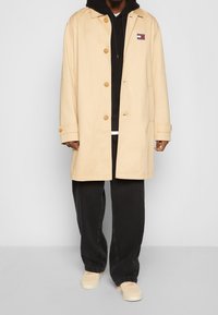 Beige lång kappa i bomull med knäppning fram, två sidofickor och en liten logotyp på vänster bröst. Bärs över en svart huvtröja.