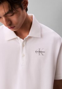 Νέος άνδρας κοιτάζει προς τα κάτω, φοράει ένα λευκό πόλο Calvin Klein με γιακά και κουμπιά, λογότυπο στο αριστερό μέρος του στήθους.