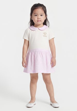 Jeune fille debout portant une robe blanche et rose clair avec un col Claudine et des ballerines blanches sur fond blanc.