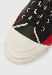 Čierna a červená teniska s textúrovaným koženým zvrškom, bielou gumovou špičkou a čiernymi šnúrkami. Má ošúchané detaily a strieborné očká.