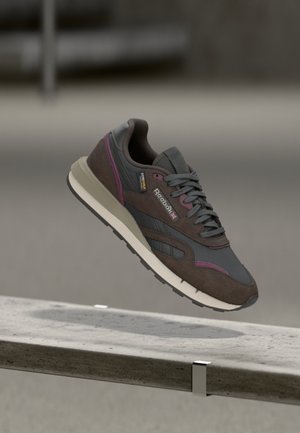 Hnedé a šedé Reebok tenisky s purpurovými detailmi plávajúce nad betónovým okrajom v neutrálnom vnútornom priestore.