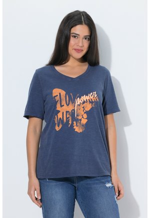 Kvinde med langt mørkt hår iført en mørkeblå V-hals T-shirt med orange "Flower Power" tekst og blomster grafik, kombineret med revnede blå jeans.