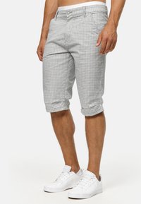 Homme portant un short gris à carreaux jusqu’aux genoux et des baskets blanches, debout devant un fond uni.