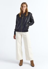 Sort læderbomberjakke med glat tekstur, guld lynlås og ribbet manchetter, kombineret med cremehvide bukser med vide ben og brune ankelstøvler.