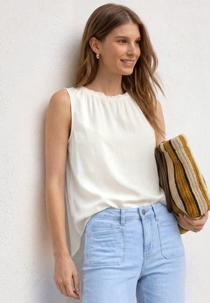 Jeune femme souriante, portant un haut blanc sans manches, un jean bleu clair, tenant une pochette tricotée rayée contre un mur blanc.