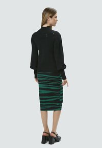 Schwarzer Strickpullover mit hohem Kragen, kombiniert mit einem grün-schwarz gestreiften Bleistiftrock. Das Outfit umfasst klobige schwarze Absätze.