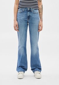 Személy fakó kék trapéznadrágot, fehér tornacipőt és tengerészkék-fehér csíkos ujjatlan felsőt visel, látható tetovált karokkal.