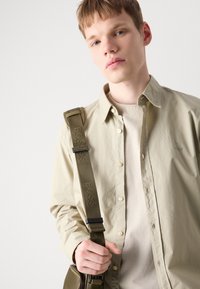 Camisa de botones de color beige claro, con una textura suave, llevada sobre una camiseta color crema. Accesorio: correa verde oliva con la marca "BOSS", sosteniendo un bolso.