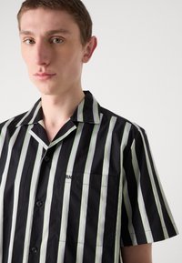 Chemise noire à manches courtes avec des rayures verticales vertes et blanches, design à col, présentant une poche poitrine et un logo de marque.