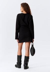 Sort langærmet mini kjole med en tætsiddende silhuet. Bæres med knæhøje sorte støvler og holder en lille sort håndtaske. Glat stofstruktur.
