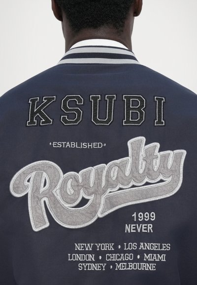 Jachetă bomber marină cu text brodat "KSUBI" și "Royalty" în argintiu, cu numele unor orașe suplimentare. Detaliu cu dungi pe guler.