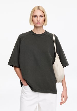 Blonde vrouw met kort haar, gekleed in een oversized donkergrijs T-shirt, witte broek, en met een kleine witte schoudertas, voor een witte achtergrond.