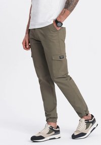Khaki cargo kalhoty vyrobené z bavlněné směsi, s elastickými manžetami, bočními kapsami a značkovou nášivkou. V kombinaci s černými a béžovými teniskami.