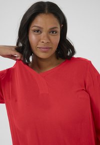 Blusa rossa a maniche corte con scollatura a V e un piccolo dettaglio a tab. Il tessuto ha una texture sottile, creando un aspetto leggero e informale.