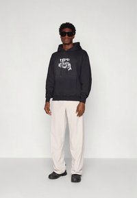 Sweatshirt preta com capuz, estampa gráfica, bolso frontal e cordão ajustável. Combinada com calças bege claro e sapatos pretos. Modelo usa óculos escuros.