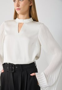 White blouse with a high neck and keyhole cutout, featuring long pleated sleeves. Paired with black high-waisted trousers and a belted waist.