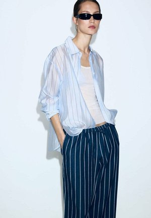 Femme portant des lunettes de soleil noires, une chemise légère rayée bleu clair par-dessus un top blanc, et un pantalon rayé bleu marine avec les mains dans les poches.