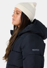 Mørkeblå dunjakke med hætte, der har et logo på brystet. Sættes sammen med en lys beige strikbeanie. Glat, struktureret stof.