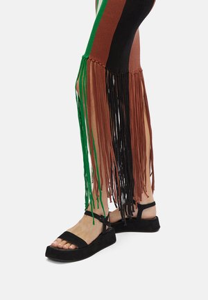 Viacfarebné, pletené legíny s strapcami; farby zahŕňajú zelenú, hnedú, čiernu a červenú. Čierne platformové sandále s popruhmi okolo členkov.