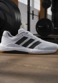 Chaussure de sport blanche avec des bandes noires et une semelle en gomme sur le sol de la salle de sport, avec des poids et une barre à l'arrière-plan flou.