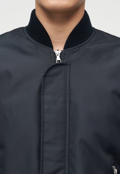 Κοντινό πλάνο ενός ατόμου που φοράει σκούρο μπλε μπουφάν-βομβιστή με ριμπ ύφασμα στον γιακά και ασημένιο φερμουάρ που είναι μερικώς κλειστό.