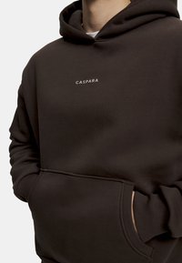 Mørkebrun hettegenser laget av myk stoff, med kengurulomme og et frontlogo i lys farge. Designet er avslappet med ribbestrikkede mansjetter.