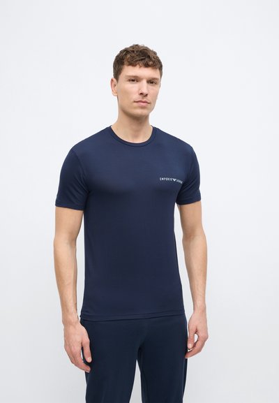 Άνδρας με κοντά σγουρά μαλλιά που φοράει μπλε ναυτικό μπλουζάκι Emporio Armani και αντίστοιχο παντελόνι, μπροστά σε απλό λευκό φόντο.