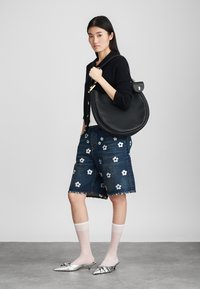 Black leather shoulder bag with a rounded shape, golden hardware, worn with a black sweater, floral-embroidered denim shorts, and metallic flats.