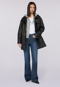 Giubbotto di pelle nera sopra un felpa grigia con cappuccio, t-shirt bianca con grafica e jeans azzurri svasati. Presenta scarpe nere a punta e capelli neri lisci.