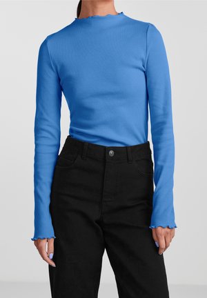 Femme portant un haut bleu côtelé ajusté à manches longues avec des bords ondulés, associé à un pantalon noir taille haute sur un fond uni.